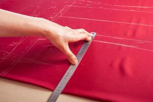 Seamstress at work. Dressmaker making clothes in modern studio. Tailor holding pencil and marking fabric. Woman standing at table with cut textile, sewing machine, thread, pins, needles, tape, cutouts photo