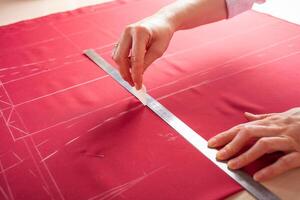 Seamstress at work. Dressmaker making clothes in modern studio. Tailor holding pencil and marking fabric. Woman standing at table with cut textile, sewing machine, thread, pins, needles, tape, cutouts photo