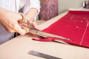 costurera a trabajar. modista haciendo ropa en moderno estudio. Sastre participación lápiz y calificación tela. mujer en pie a mesa con cortar textil, de coser máquina, hilo, patas, agujas, cinta, recortes foto