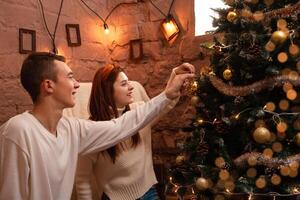 Young people in love hang a toy on the Christmas tree. A guy and a girl dress up a Christmas tree. New Year's decorations in the photo studio. Posing for models.