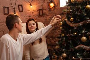 Young people in love hang a toy on the Christmas tree. A guy and a girl dress up a Christmas tree. New Year's decorations in the photo studio. Posing for models.