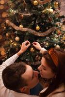 Young people in love hang a toy on the Christmas tree. A guy and a girl dress up a Christmas tree. New Year's decorations in the photo studio. Posing for models.