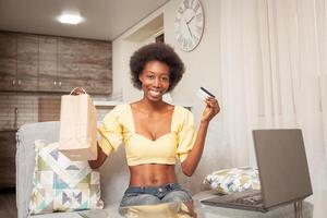 africano americano negro mujer a hogar sentado en frente de computadora portátil, en línea compras, hogar entrega, banco tarjeta. descuentos promociones en el almacenar. contento sonrisas foto