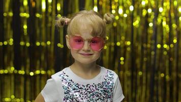 kind lachend, op zoek Bij camera. meisje in roze zonnebril poseren Aan achtergrond met folie gordijn video