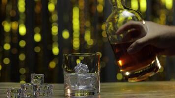 gieten van gouden whisky, cognac of brandewijn van fles in glas met ijs kubussen. glimmend achtergrond video