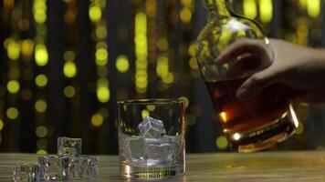 Pouring of golden whiskey, cognac or brandy from bottle into glass with ice cubes. Shiny background video