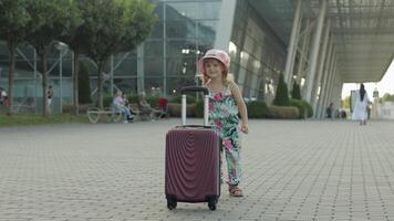 bambino ragazza turista con valigia Borsa vicino aeroporto. ragazzo balli, gioisce, celebra con bagaglio video