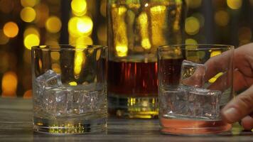 Pouring of whiskey, cognac or brandy from bottle into glasses with ice cubes. Shiny background video