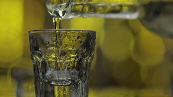 Barman pour frozen vodka from bottle into shot glass against shiny gold party background. Close up video