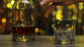 Pouring of golden whiskey, cognac or brandy from bottle into glass with ice cubes. Shiny background video