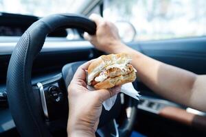 Asian woman driver hold and eat hamburger in car, dangerous and risk an accident. photo