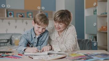 grootmoeder en kleinzoon zijn aan het doen huiswerk. portret van een gelukkig familie. aan het leren Bij huis. onderwijs. grootmoeder en kleinzoon lachen. schattig emoties in de kader. de verhaal van een familie. video