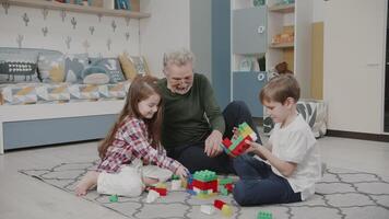 grand-père pièces avec le sien petits enfants dans jouets. un vieux homme, une petite fille et une petit fils sont bâtiment une constructeur. mignonne des gamins sont souriant et en riant dans le garderie. le récit de un content famille. video