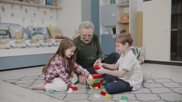 Opa Theaterstücke mit seine Enkelkinder im Spielzeuge. ein alt Mann, ein Enkelin und ein Enkel sind Gebäude ein Konstrukteur. süß Kinder sind lächelnd und Lachen im das Kindergarten. das Geschichte von einer glücklich Familie. video