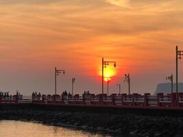 Cityscape Embraced by the Warm Glow of Sunset with Silhouetted Towers, Industrial Structures, and a Serene Pier by the Water, as the Sky Paints a Palette of Orange Hues in the Early Evening Light photo