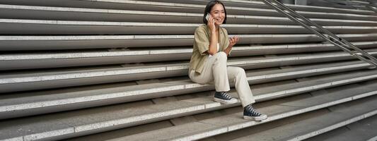 Smiling asian girl sits on stairs of building and talks on mobile phone, relaxing during telephone conversation photo