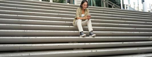 Portrait of smiling asian girl sits on stairs outdoors, sending message, using smartphone app, looking at mobile phone screen photo