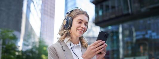 retrato de sonriente joven mujer de negocios escuchando música en auriculares y utilizando móvil teléfono mientras en un ocupado ciudad calle foto