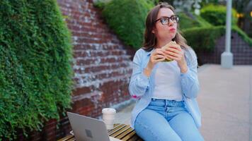 mulher tendo uma almoço com uma sanduíche e café dentro a parque com uma computador portátil video