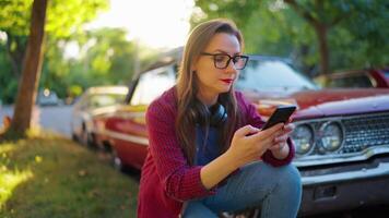 mulher é agachado baixa Próximo a vermelho vintage carro e usando Smartphone video