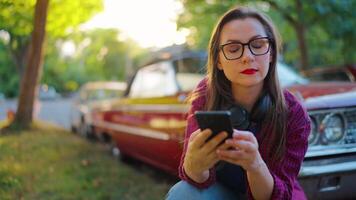 mujer es agachado abajo siguiente el rojo Clásico coche y utilizando teléfono inteligente video