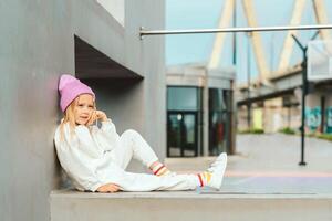 beautiful blonde girl in a white tracksuit sits on a concrete fence against the backdrop of a city landscape, copy space photo