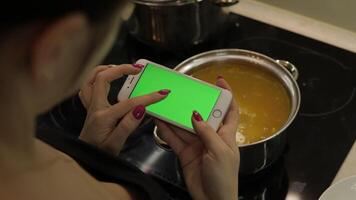 Woman is holding a smartphone with a green screen, and cooking soup video