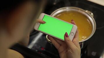 Woman is holding a smartphone with a green screen, and cooking soup video