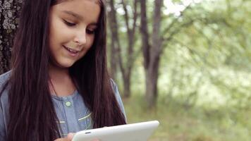 Portrait. A beautiful girl with long black hair in a dress is standing by a tree in the nature using a tablet and looking at something interesting in it and laughing video