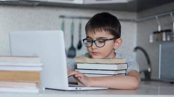 A schoolboy in glasses does homework while sitting in the kitchen at the table using a lot of textbooks and a laptop. Online learning video