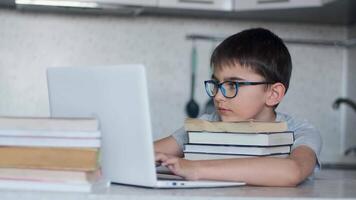 Attractive schoolboy with glasses doing homework while sitting in the kitchen at the table using a lot of textbooks and a laptop. Online learning video