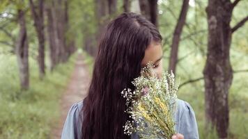 en trevlig skön liten flicka med lång hår är stående förbi en träd i ljus solig väder och sniffa en bukett av blommor video