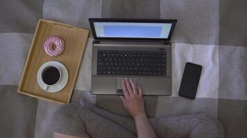 Overhead shot, creative girl working using a dark color laptop and a mobile phone sitting on the bed at home and drinking a coffee with donut. 4K video