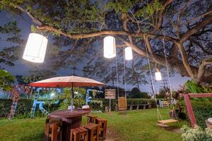 grande lluvia árbol decorativo Encendiendo arte lámpara con de madera mesa silla en césped en restaurante foto