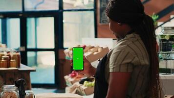 Small business owner looking at phone with greenscreen in local grocery store, showing blank copyspace layout on smartphone. Vendor selling her homegrown produce at farmers market. video