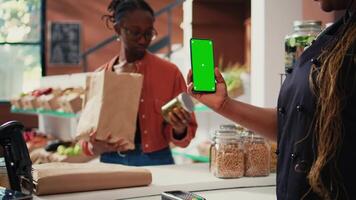 Store vendor holding phone with greenscreen display, working at cash register to serve customers. African american seller using isolated mockup template on smartphone screen. Tripod shot. video