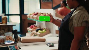 African american vendor using greenscreen template at counter, presenting chromakey display on smartphone. Woman seller holding phone with isolated mockup layout and copyspace. video