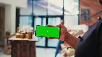 Local merchant showing greenscreen layout on phone, holding device with isolated copyspace template at cash register. African american retailer with smartphone running chromakey display. Tripod shot. video