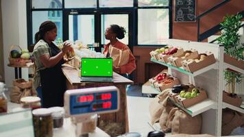 Seller recommending products next to greenscreen on laptop, presenting organic bulk items in local grocery store. Computer placed on stall showing blank mockup isolated template. Tripod shot. video