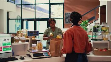 Merchant weighting bulk items on scale for regular client, selling various types on homemade products in reusable jars at checkout. Vendor chatting with buyer about healthy eating. Handheld shot. video