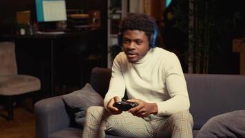 Gamer wearing headphones, feeling happy after receiving winner screen on gaming console for killing all enemies. African american player at home bragging after being victorious in videogame video