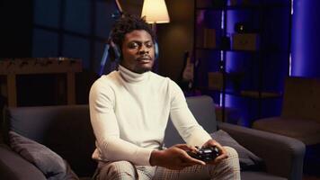 BIPOC man playing shooter videogame in neon lit apartment, holding controller. Gamer participating in internet multiplayer game using console system and headphones, zoom out shot video