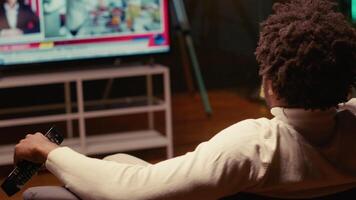 Man holding remote, watching news channel on television broadcast. Focus on person relaxing on couch in apartment by looking at widescreen smart TV set in blurry background video