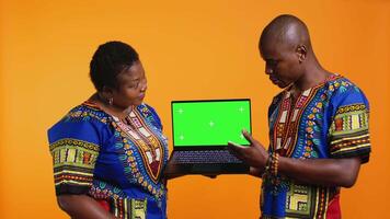 Ethnic people presenting laptop with greenscreen layout, showing blank mockup chromakey display and posing in studio. African american husband and wife pointing at isolated copyspace. video
