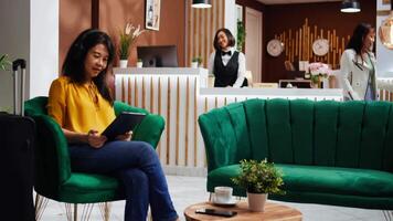 Asian woman checking webpages on tablet in hotel lobby, waiting to see accommodation and start holiday. Tourist relaxing in lounge area after long flight, using device for internet browsing. video