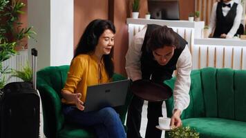 Asian woman receiving coffee while she works on laptop, sitting in lounge area at five star hotel and waiting for registration. Waiter serving drinks for guest relaxing, hospitality industry. video