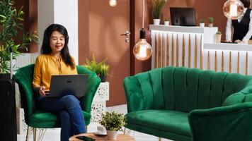 Asian tourist ordering coffee cup while she works on laptop, waiting for check in process in lounge area lobby. Woman arriving early at hotel, relaxing in foyer with refreshment. video