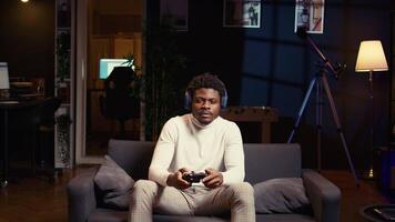 African american man playing shooter videogame in warm apartment, holding controller. Gamer participating in PvP online multiplayer game using console system and headphones for immersive experience video