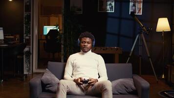 BIPOC man using headphones mic to discuss strategies with teammates while playing videogame in cozy apartment. Player chatting with online strangers while enjoying game on gaming console video