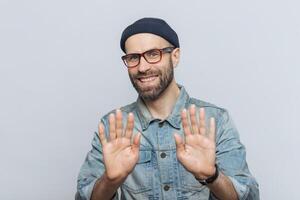 Happy man in glasses and denim jacket showing his palms to the camera, with a friendly stop gesture, wearing a beanie, on a gray backdrop photo